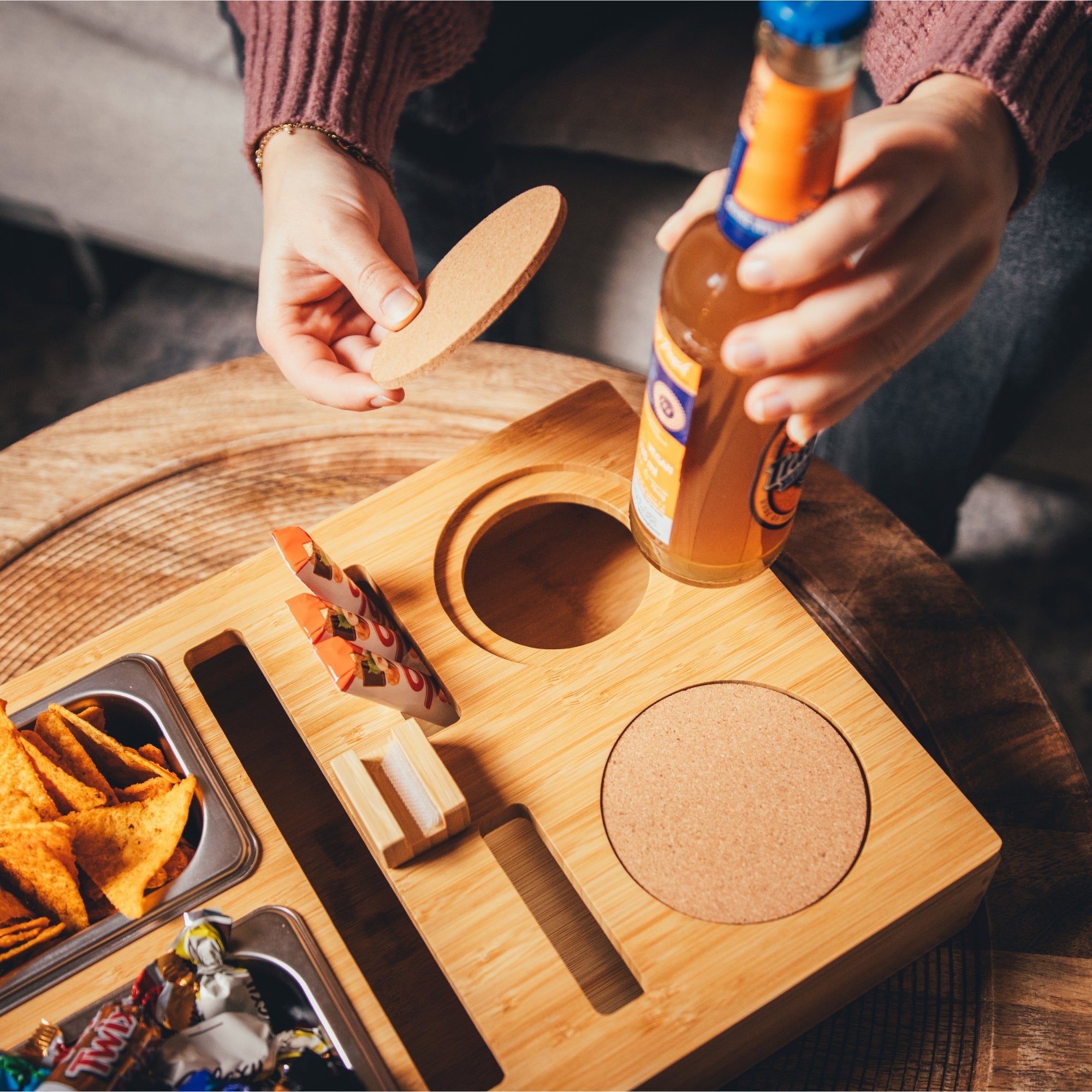 Personalisierbare Couchbar aus Holz mit Getränkehalter, runden Kork-Untersetzern und integrierten Snackfächern, gefüllt mit Chips und Süßigkeiten, ideal für gemütliche Abende auf dem Sofa
