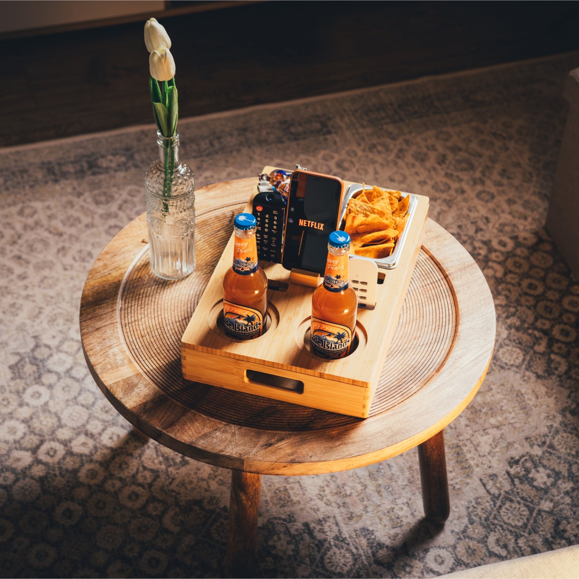 Personalisierbare Couchbar aus Holz mit integrierten Getränkehaltern, Snackbox und Smartphone-Halterung, stilvoll auf einem Beistelltisch platziert, ideal für gemütliche Fernsehabende im Wohnzimmer, von das-brettchen.de
