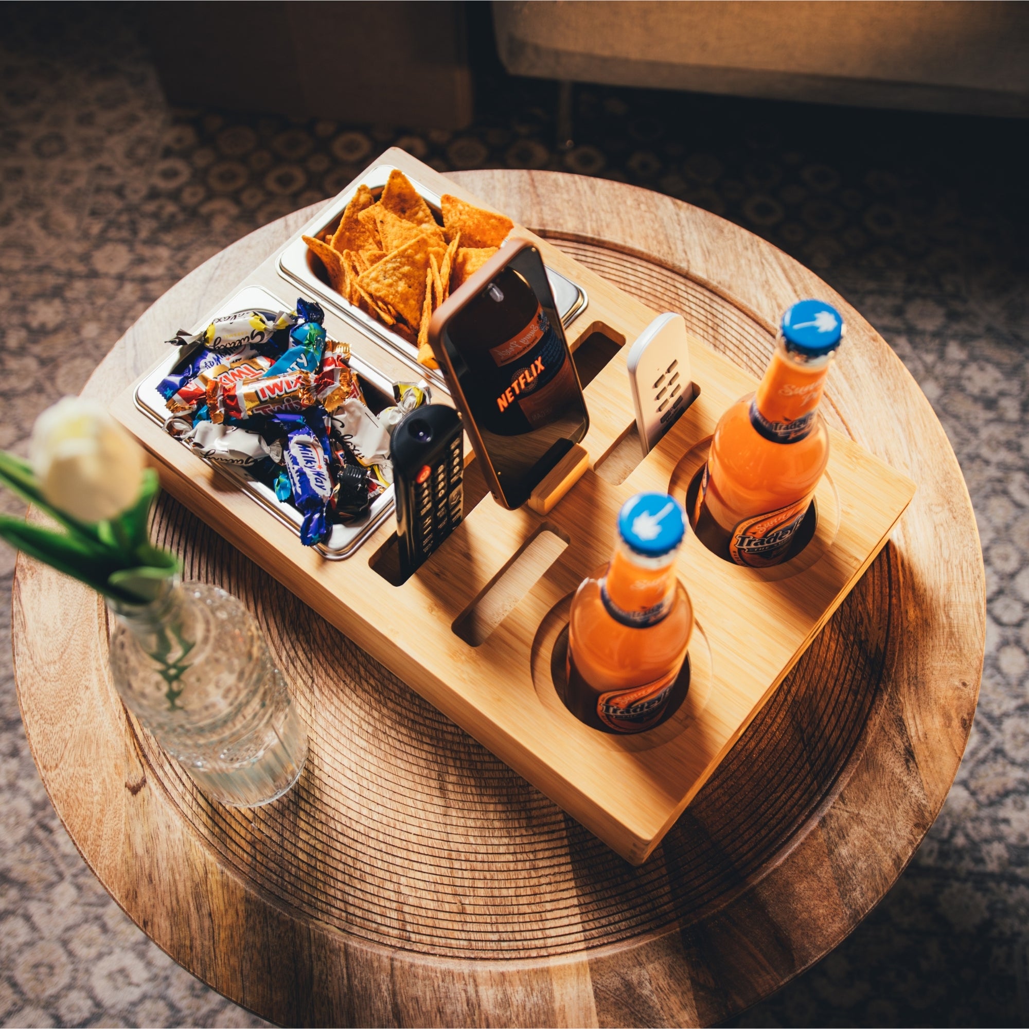 Personalisierbare Couchbar aus Holz mit integrierten Getränkehaltern, Snackfächern und Smartphone-Halterung, gefüllt mit Chips, Schokoriegeln und Getränken, auf einem runden Beistelltisch platziert, perfekt für gemütliche Fernsehabende, von das-brettchen.de